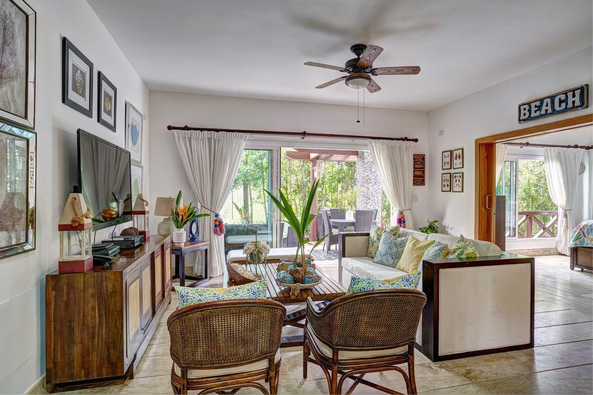 The spacious living area features a comfortable seating arrangement with wicker chairs and a sofa, complemented by light curtains. A flat-screen television is positioned against a decorative wall. Large sliding doors provide access to the outdoor patio, inviting natural light into the room.