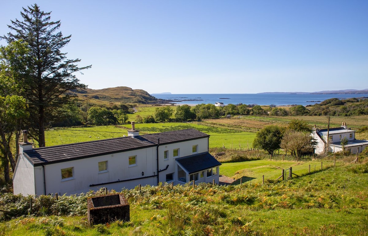 A view of the fully renovated croft house, showcasing its white exterior against a backdrop of green fields and the blue sea. The landscape features rolling hills and the distant shore, capturing the tranquil surroundings of the property.
