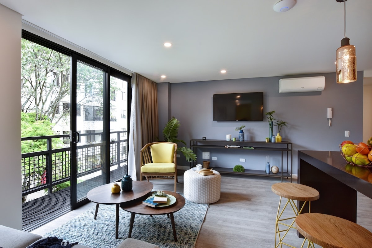 A modern living area is shown, featuring a yellow accent chair and a coffee table with decorative items. Large sliding glass doors open to a balcony, allowing natural light to fill the space. A minimalist console holds a smart TV, and a vibrant rug adds texture to the floor.