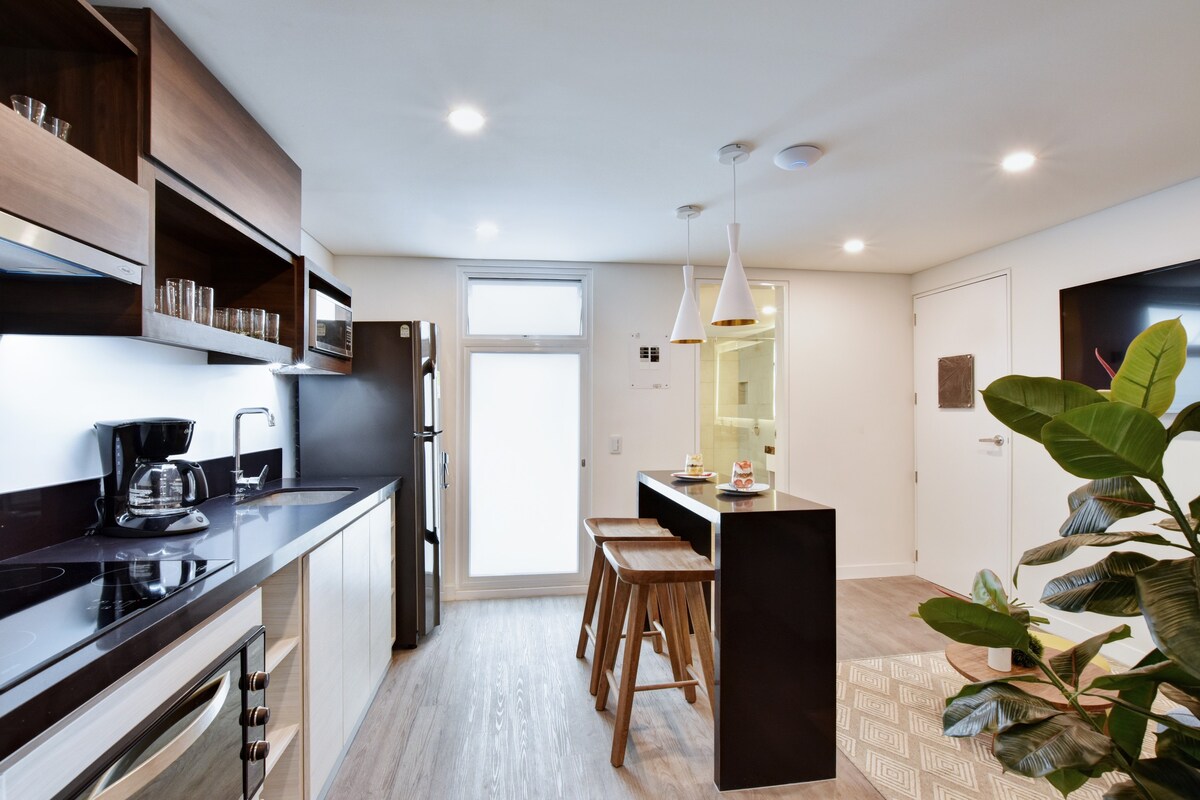 A modern kitchen showcases sleek cabinetry and stainless steel appliances. A coffee maker, along with glassware, is neatly arranged on the counter. Two wooden stools provide seating at the breakfast bar, while a potted plant adds a touch of greenery.