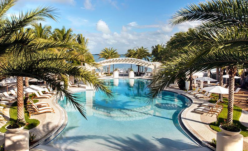 A spacious pool area is surrounded by tropical palm trees, providing a serene atmosphere. Sun loungers are arranged neatly around the pool, with gentle waves reflecting the blue sky. An elegant white pergola adds a touch of sophistication to this inviting outdoor oasis.