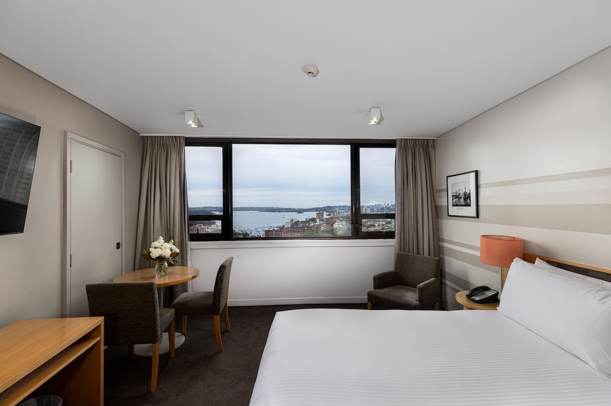 A room features a spacious king-size bed, accompanied by a small dining table with two chairs. Large windows offer expansive views of the water and city, while modern decor complements the inviting ambiance. A comfortable armchair sits beside the table.