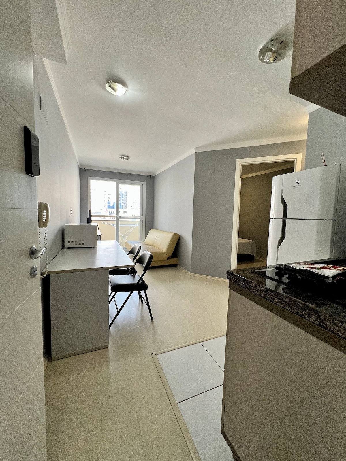 A compact living space is shown, featuring a small dining area with a table and two chairs. A sleek kitchen countertop is visible on the right, and a convertible sofa is positioned along the wall. Large windows provide natural light and a view of the outside.