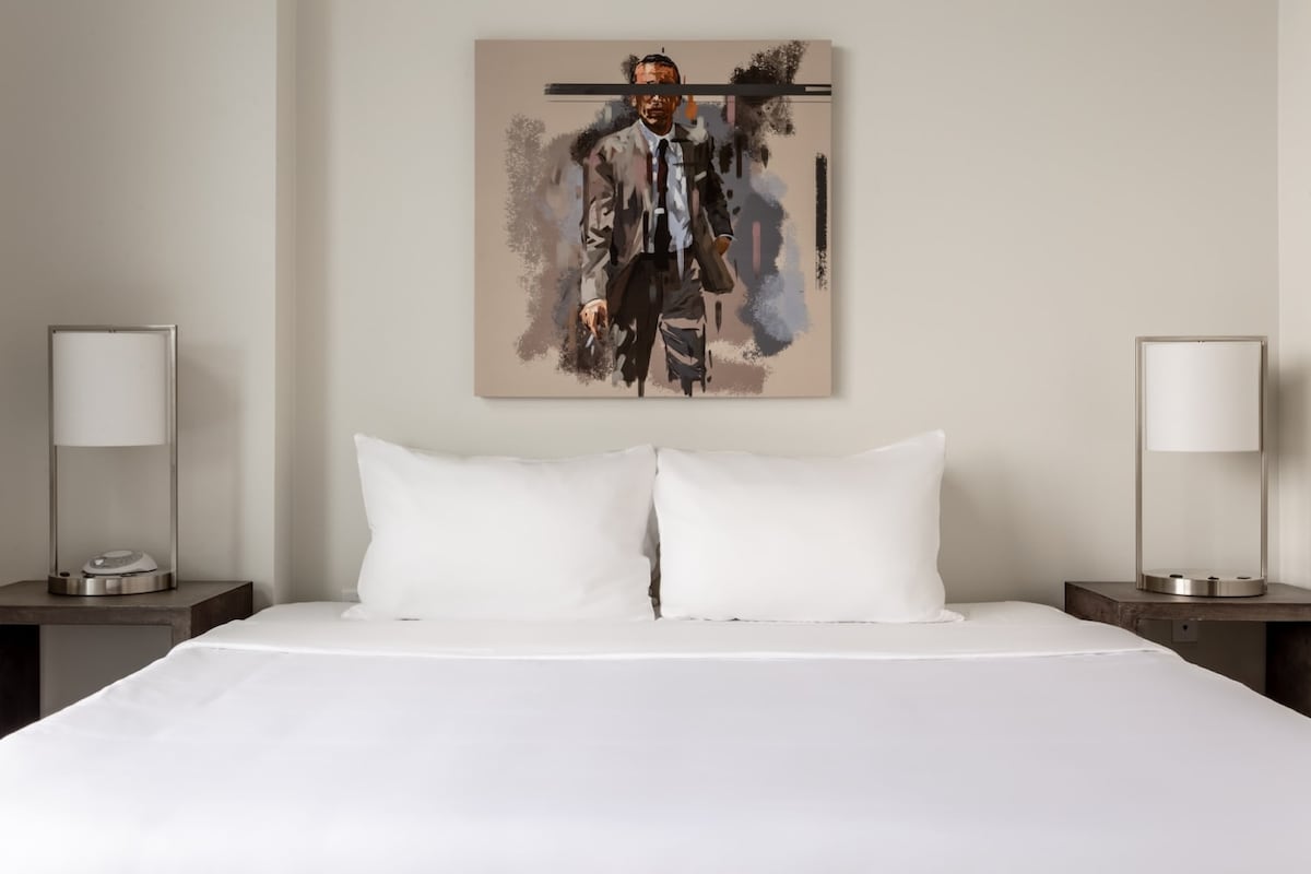 A spacious bed with two plush white pillows is centered against a neutral wall. On either side, modern nightstands hold stylish lamps. Above the bed, a contemporary art piece featuring a blurred figure adds a touch of sophistication to the room.