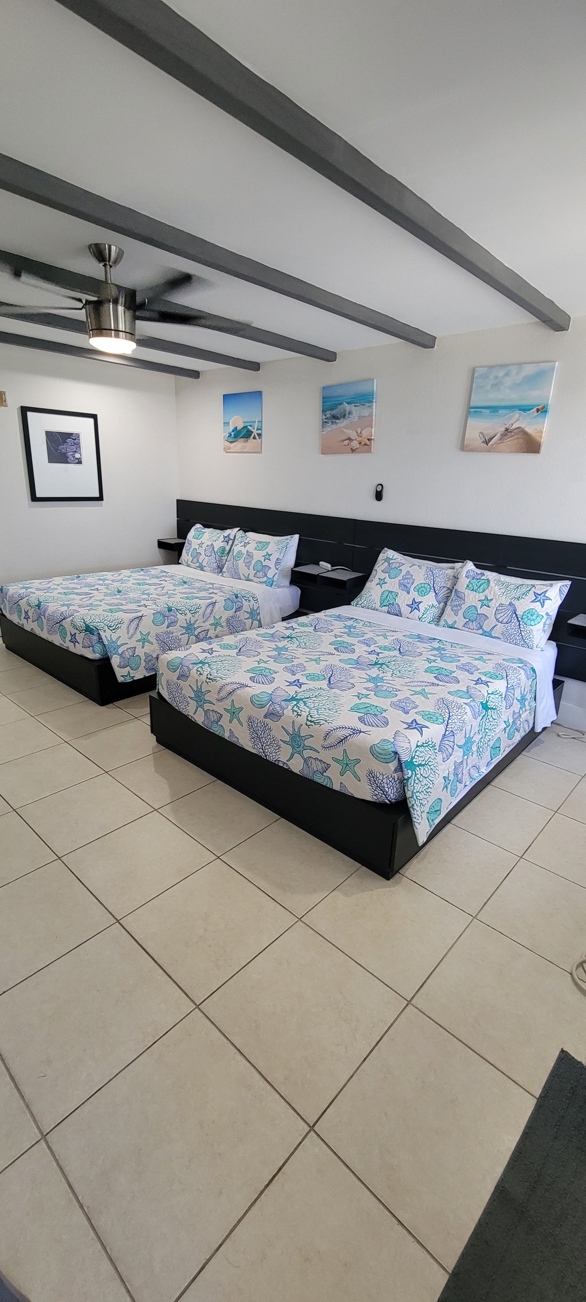 The spacious studio contains two luxurious full beds, each adorned with light blue and green island-inspired bedding. Light-colored tile flooring enhances the bright atmosphere, complemented by ceiling beams. Ocean-themed artwork decorates the walls, providing a relaxed coastal feel.