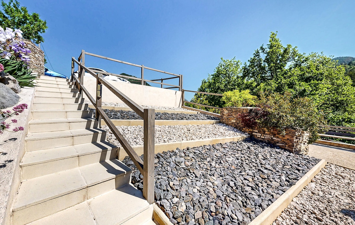 A well-maintained outdoor staircase is surrounded by decorative stone landscaping. The steps are flanked by wooden railings, leading to a pathway that rises gently toward a scenic area adorned with trees and colorful flowers, set against a clear blue sky.