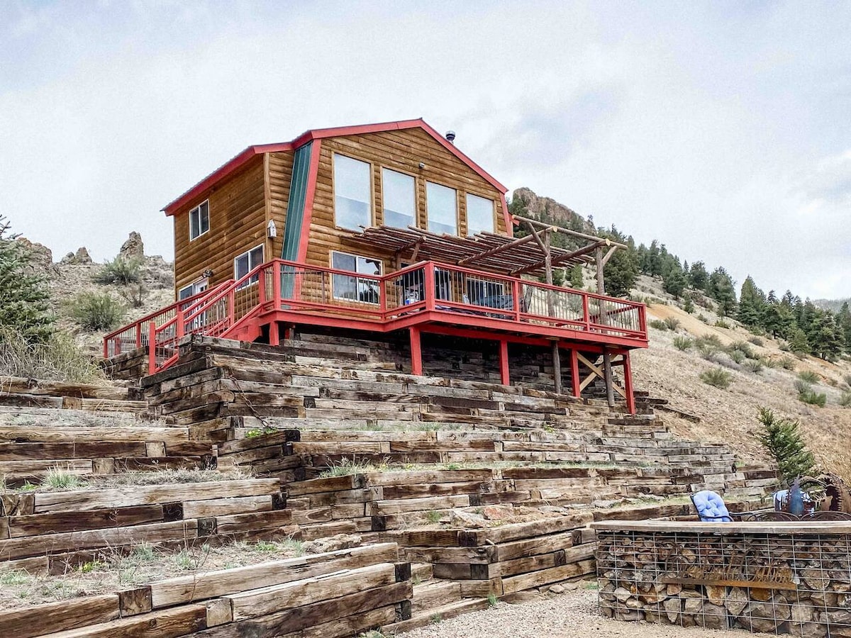 A cabin with a red and wood exterior is positioned on a hillside, showcasing a wide deck with railings. The structure has multiple large windows, allowing for natural light, while stacked timber steps lead up to the entrance, surrounded by natural landscape.