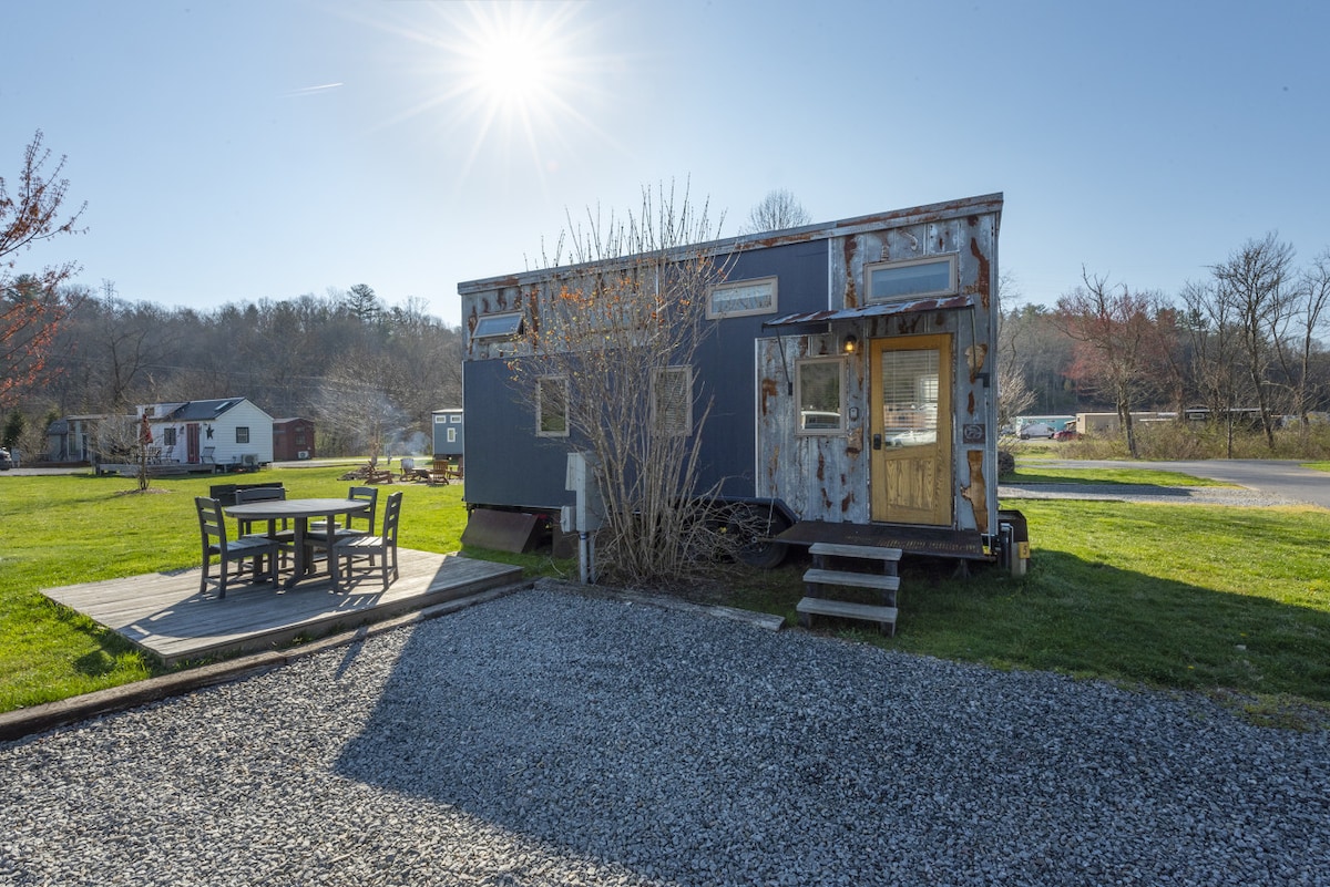 A tiny home with a weathered exterior and wooden door is positioned in a grassy area. A spacious patio accompanies the home, featuring a wooden table and chairs. The sun shines brightly in the sky, casting light on the nearby trees and greenery.