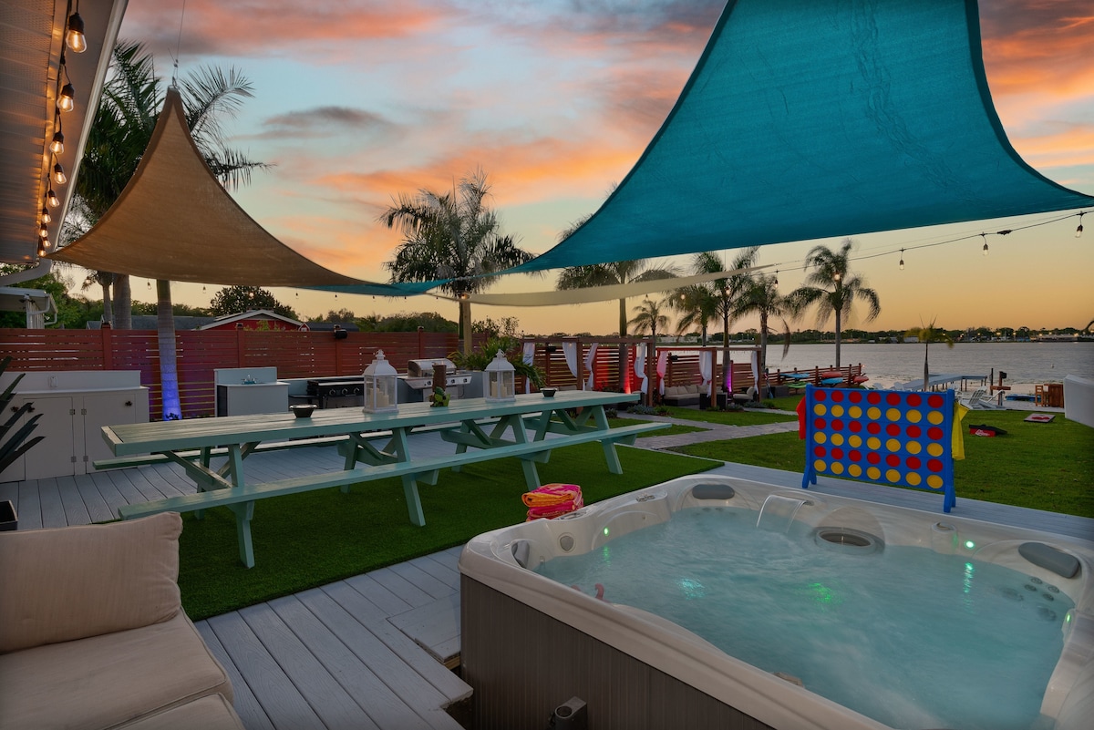 A spacious outdoor area is set against a serene lake backdrop, featuring a hot tub and dining tables under shaded canopies. Brightly colored seating and yard games are positioned throughout the tropical-inspired yard, with palm trees gently swaying in the evening light.