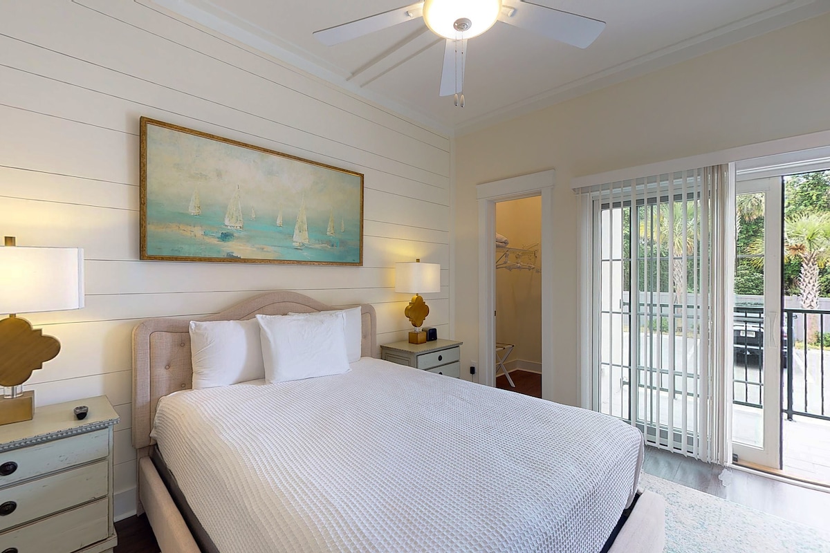The spacious bedroom features a large bed with a light-colored quilt and two bedside lamps. A piece of coastal artwork hangs above the bed. A door leads to a private balcony, and large glass doors provide natural light and a view of the outdoor area.