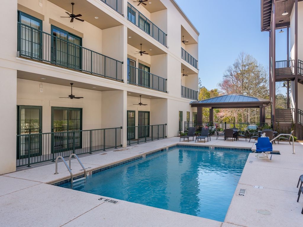 An outdoor swimming pool is set among a modern building with multiple balconies. Lounge chairs are positioned around the pool area. In the background, trees and landscaping provide a natural touch, complementing the contemporary architecture.