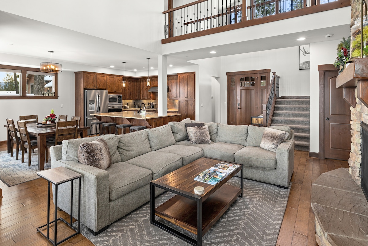 A spacious living area features a large sectional sofa arranged around a coffee table. Wooden accents and a stone fireplace create a cozy ambiance. The open floor plan connects to a dining area with a wooden table, while a staircase leads to the upper level, illuminated by natural light.