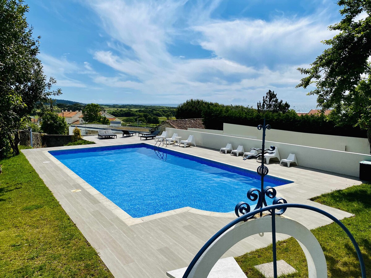 The outdoor space features a large swimming pool, surrounded by well-maintained grass and lounge chairs. A scenic view of the landscape is visible beyond the pool area, under a clear blue sky with scattered clouds.