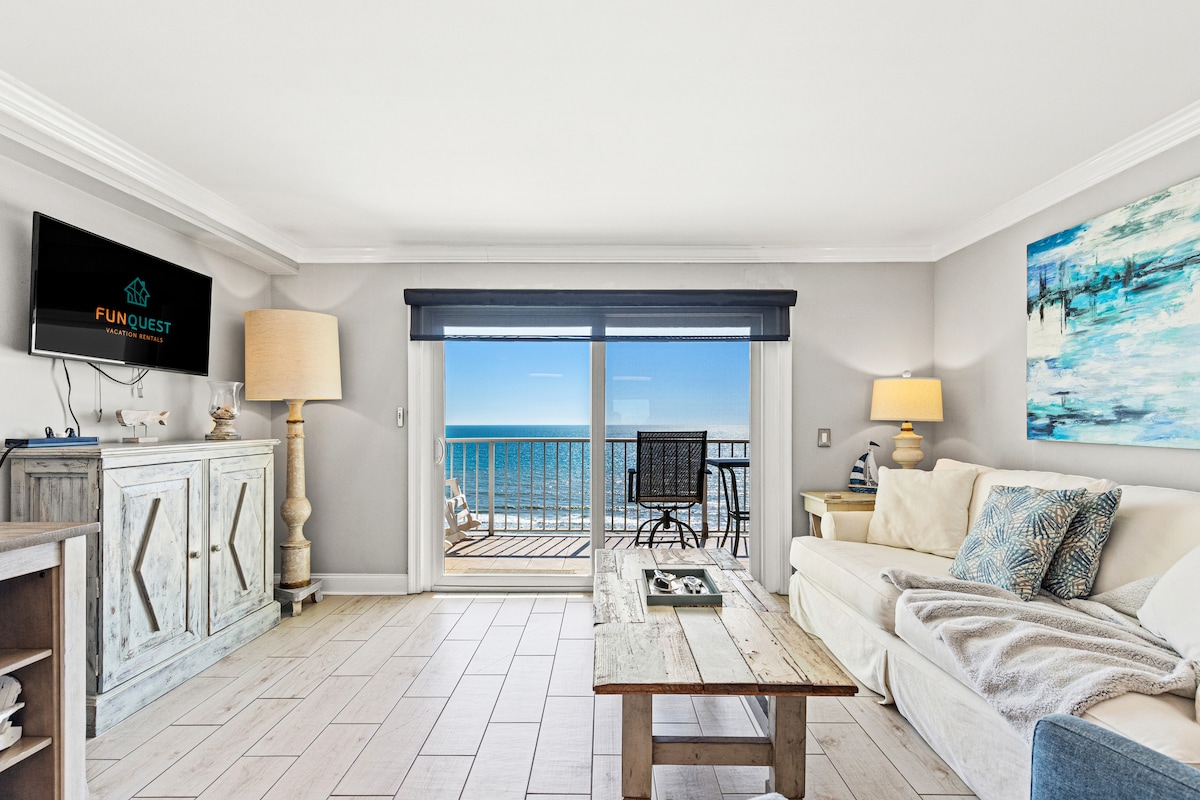 A bright and airy living space features large glass doors opening to a balcony with ocean views. A light-colored sofa is positioned beside a rustic wooden coffee table, and a television is mounted on the wall. Soft lighting from lamps creates a relaxed ambiance.