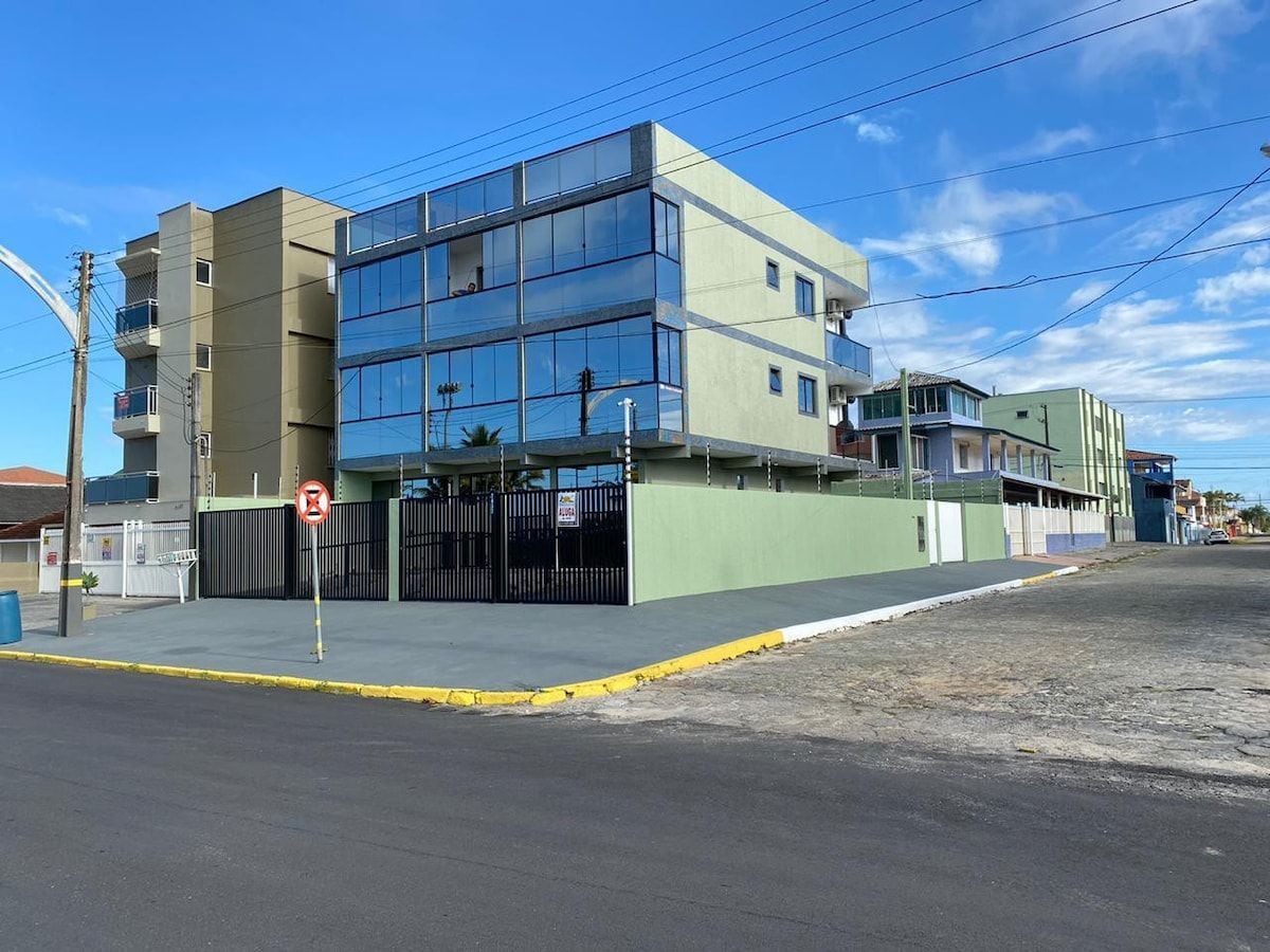 The exterior of the building is showcased, featuring a modern three-story structure with large reflective windows. A secure entrance, framed by a low fence, is visible. The paved area in front is clear of obstacles, providing easy access. Surrounding buildings and a blue sky enhance the scene.