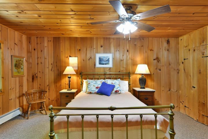 किटी फ़ॉय हाउस में एक देहाती बेडरूम, जिसमें दीवारों और छत पर पाइन की लकड़ी के पैनल लगे हुए हैं, जो गर्माहट का एहसास देते हैं। कमरे के बीचों-बीच पीतल के फ़्रेम वाला एक बेड है, जिस पर सफ़ेद और नीले रंग के पैटर्न वाली रज़ाई बिछी हुई है। बेड के दोनों ओर लकड़ी के नाइटस्टैंड हैं, जिन पर मैच करने वाले लैम्प रखे हुए हैं।