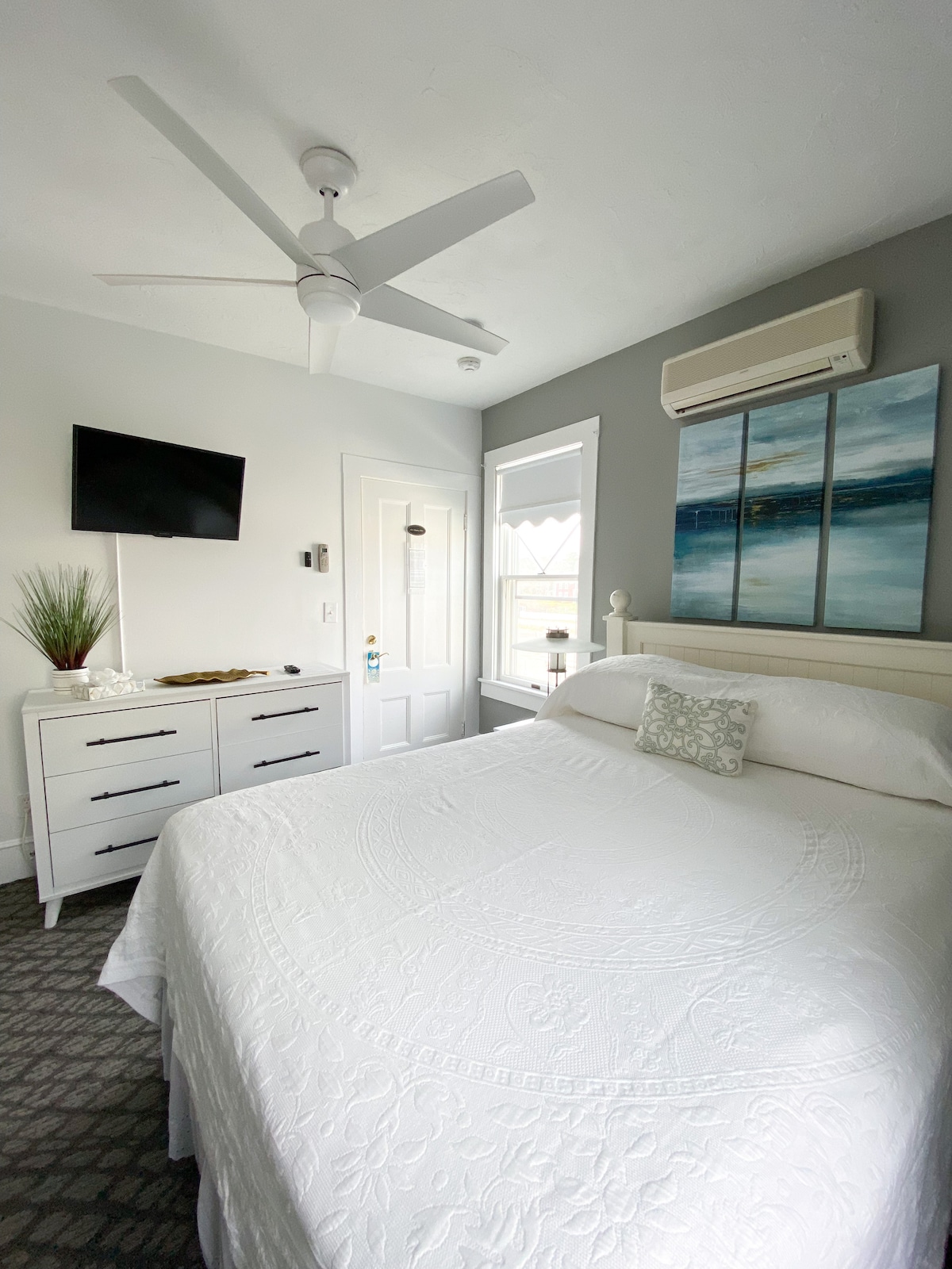 A cozy room features a queen bed with a white coverlet, complemented by decorative pillows. A flat-screen TV is mounted on the wall, and a dresser offers additional storage. Natural light streams through a window, while a ceiling fan provides air circulation.