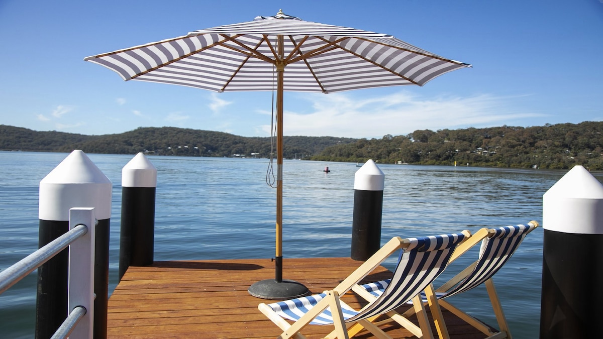 A wooden jetty extends over calm waters, featuring two striped lounge chairs positioned beneath a large, striped umbrella. White and black posts mark the edges, with rolling green hills visible in the background under a clear blue sky.
