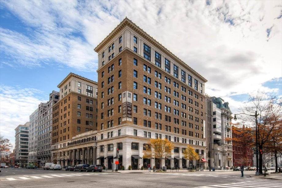 The 11-story Beaux-Arts building showcases a flat roof and arched windows, presenting a striking presence on the corner. The first story is rusticated and raised, with trees lining the sidewalk, creating a welcoming atmosphere. The sky above features scattered clouds filtering natural light.
