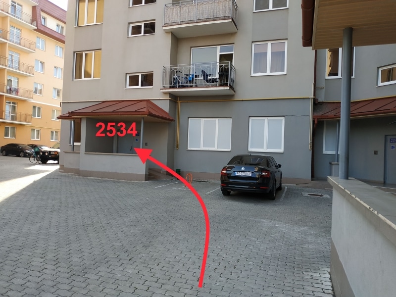 A paved parking area is visible in front of a modern apartment building. The structure features large windows and a balcony on the second floor. A marked vehicle is parked nearby, providing a glimpse of convenient onsite parking.