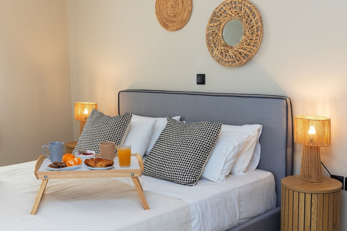 A neatly arranged queen size bed is presented with a tray holding drinks and pastries. Decorative pillows enhance the bed's comfort, while woven wall art and mirrored accents add subtle texture to the neutral walls. Soft lighting from bedside lamps provides a cozy atmosphere.