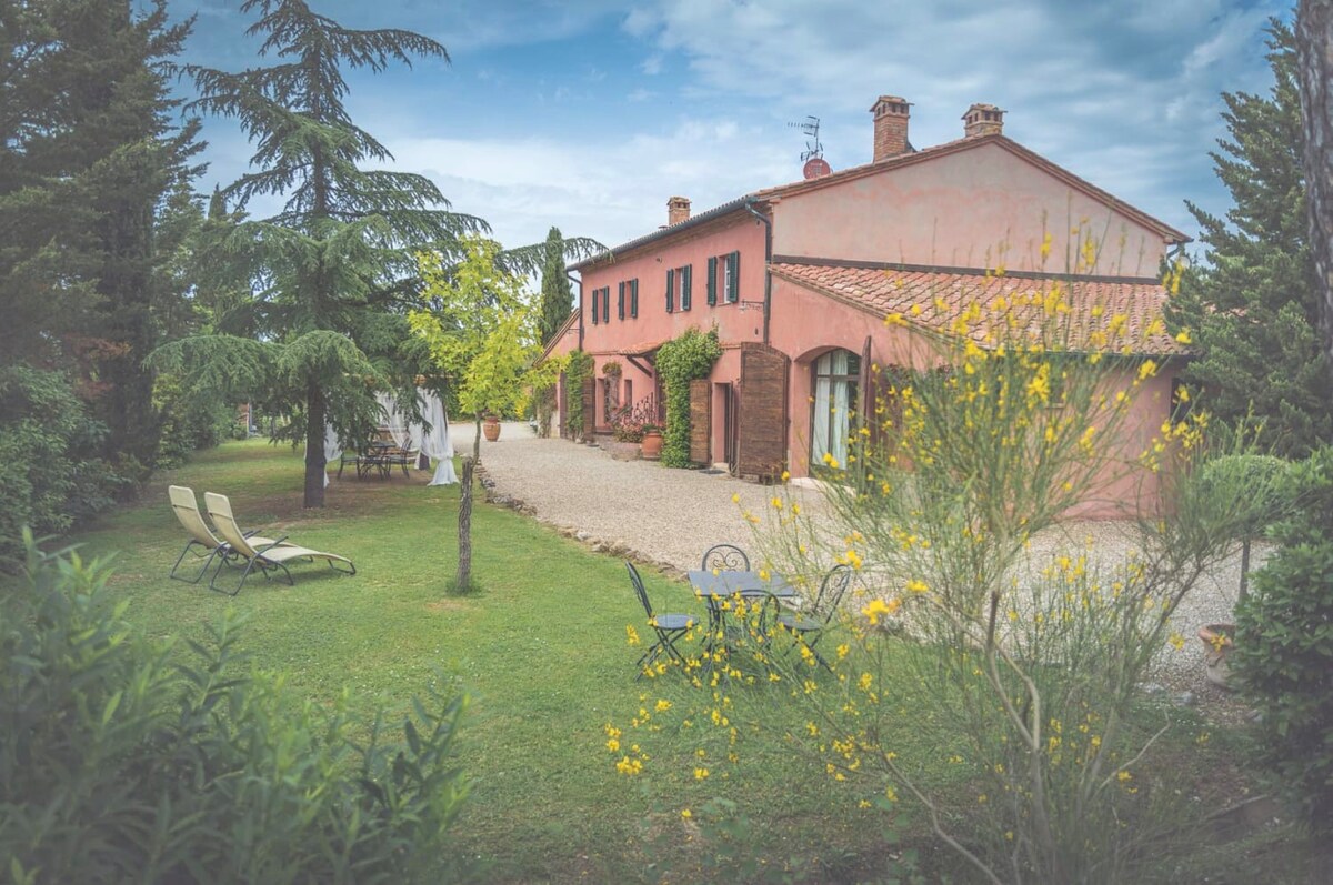 A charming farm building features a soft pink exterior with dark shutters, surrounded by a lush garden. Loungers are positioned on the grass, and a small outdoor dining set is visible. Lush greenery and blooming yellow flowers enhance the tranquil atmosphere.