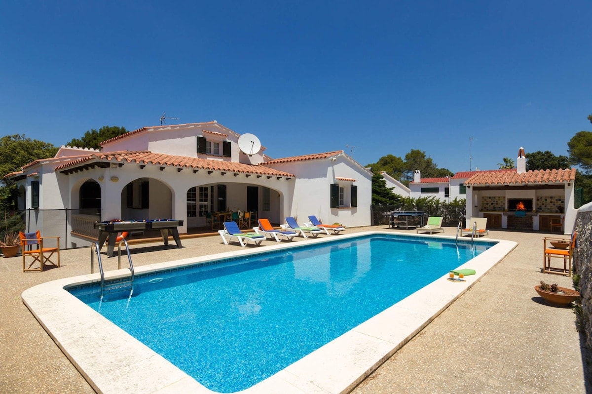 A spacious outdoor area features a sparkling private pool surrounded by lounge chairs. A barbecue stand is located nearby, and the villa's charming architecture is complemented by various trees and shrubs. Clear blue skies provide a bright backdrop to this inviting setting.