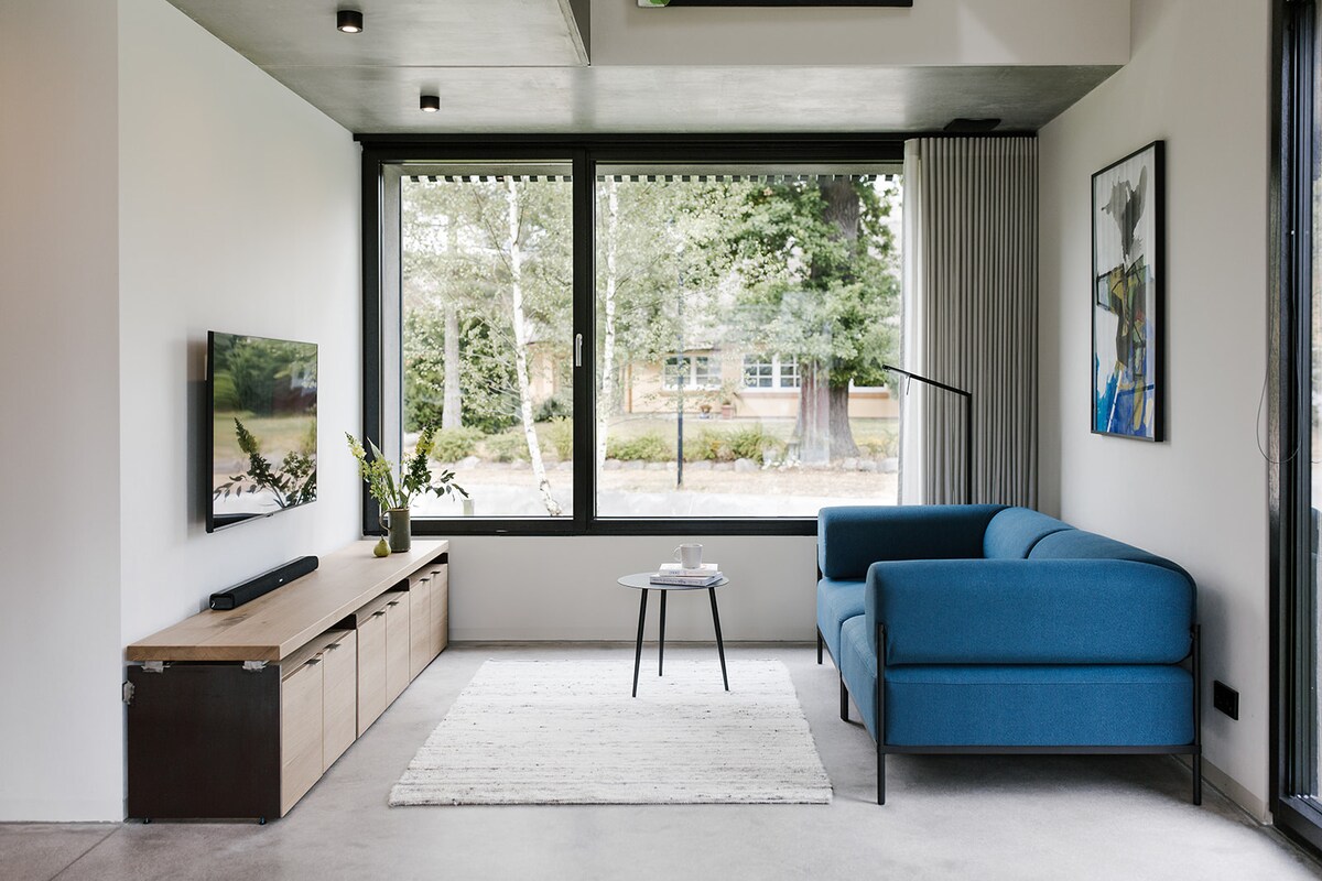 A bright living area features a modern blue sofa facing a wall-mounted television. A small coffee table sits on a light rug, complemented by a wooden sideboard. Large windows allow natural light to fill the space, overlooking a green garden.