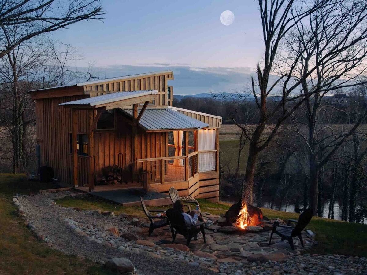 A cozy tiny house is illuminated by soft evening light, with a fire pit surrounded by several chairs. A river is visible nearby, framed by leafless trees under a clear sky featuring a rising moon.