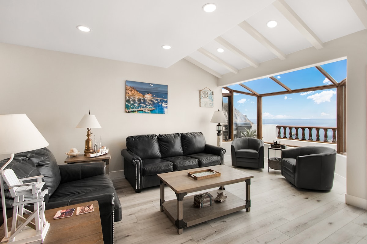 The living room features two black leather sofas and a reading chair arranged around a wooden coffee table. A panoramic window offers ocean views, while artwork depicting coastal scenery adorns the walls. Soft lighting is provided by ceiling fixtures and lamps.