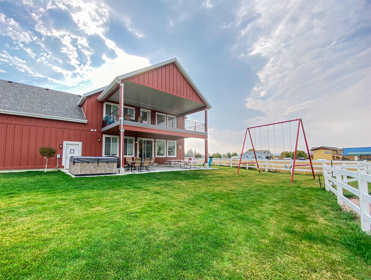 The exterior view of the farmhouse showcases a spacious grassy area with a large red building. A covered deck extends from the upper level, providing seating options. A swing set is situated on the lawn, inviting outdoor play. The sky features soft clouds, enhancing the tranquil setting.
