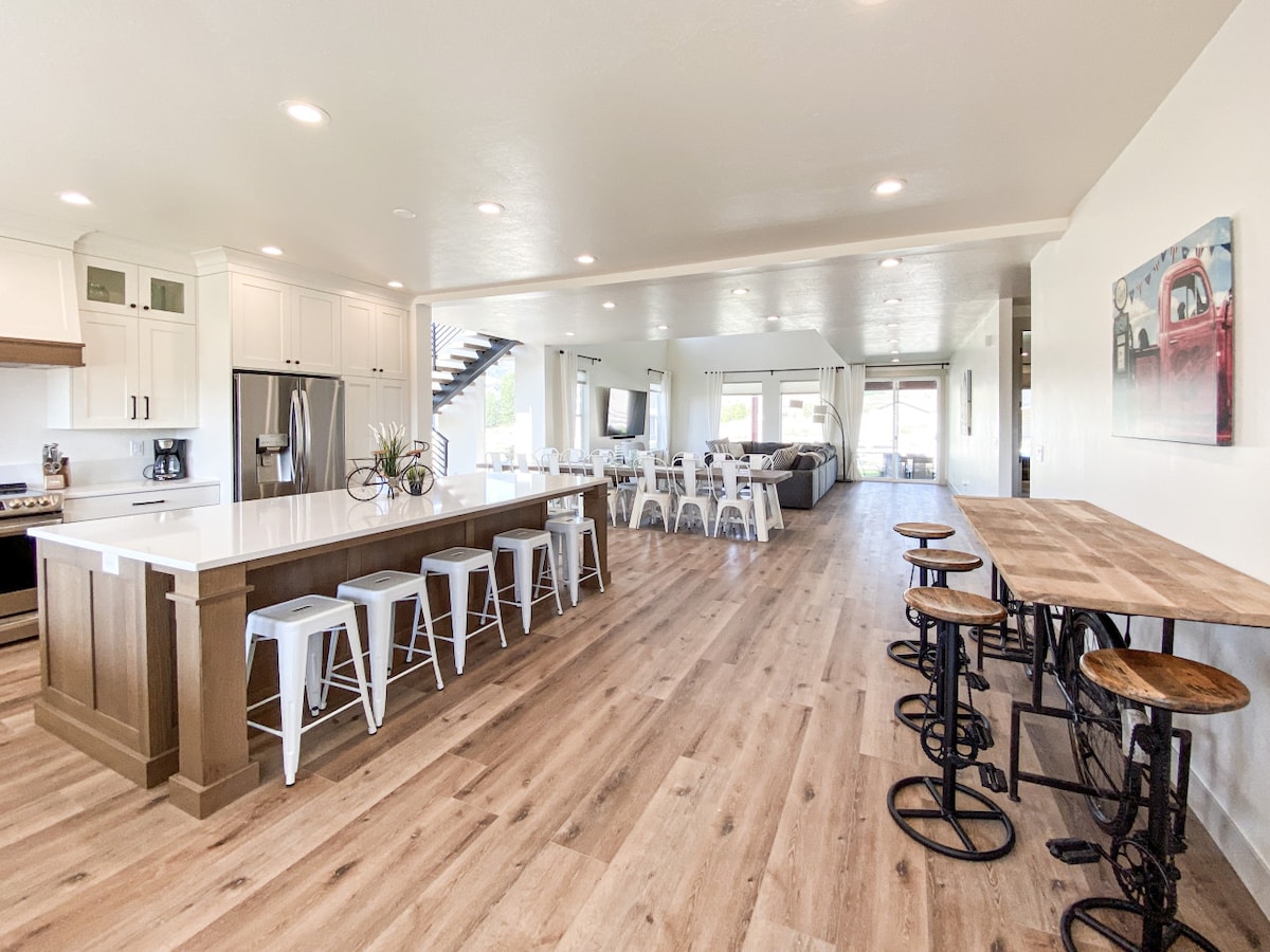 An open-concept living area features a spacious kitchen with a central island and seating for six. Metal bar stools are arranged around a wooden counter, and large windows provide ample natural light. The adjacent dining area and living space are visible in the background.