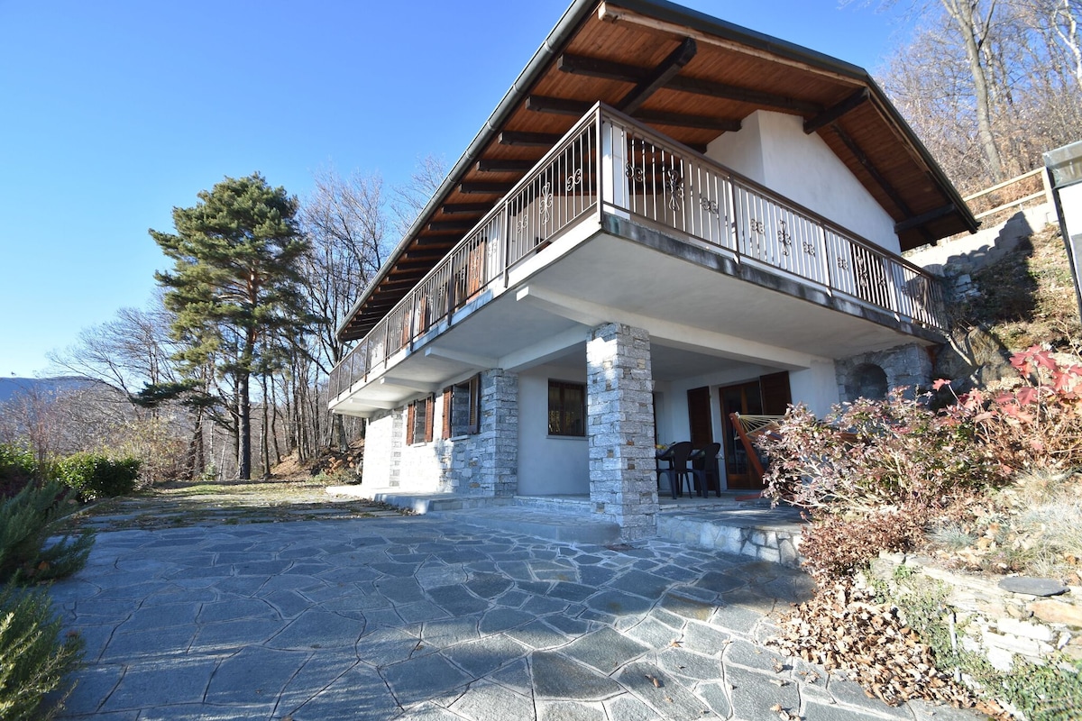 The exterior of the house is characterized by a sturdy stone foundation and a wooden balcony. Surrounding areas feature a neatly maintained garden with diverse shrubbery. A clear blue sky complements the structure, providing a serene backdrop against the natural landscape.