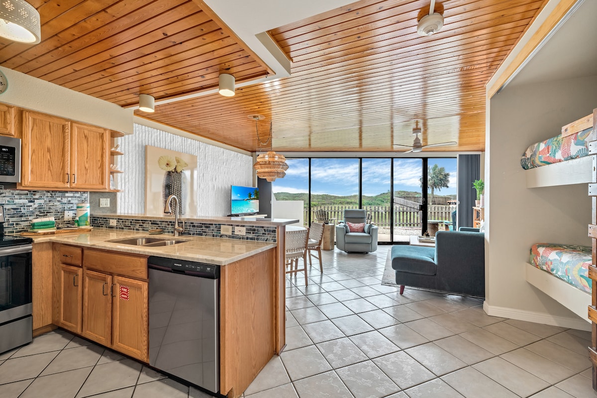 An open-concept kitchen and living area feature wooden cabinets and stainless steel appliances. A dining area with cushioned seating for four complements the tiled flooring. Large glass windows offer a view of the outdoors, while natural light illuminates the space with a warm tone.