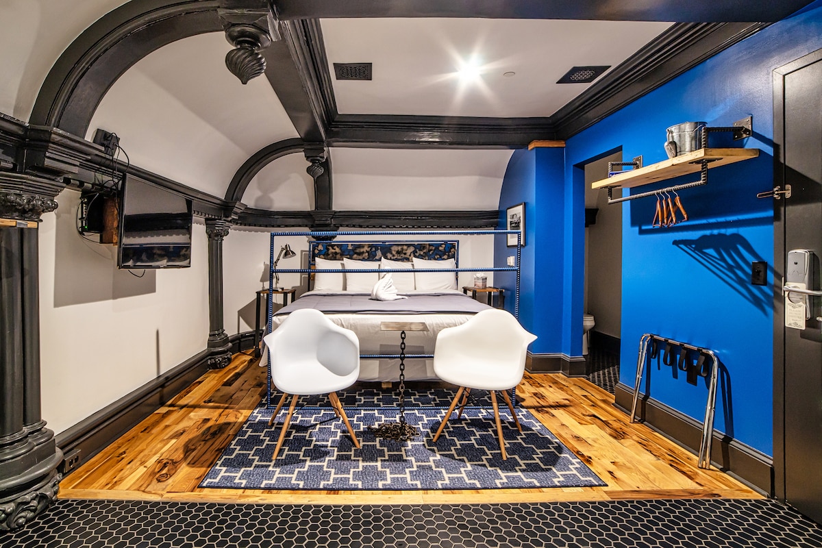 The guest room showcases a spacious bed centered against a vivid blue wall. Two white chairs are positioned on a patterned rug, enhancing the room's modern design. Industrial-style beams and elegant architectural details frame the area, while a flat-screen TV is mounted for convenience.