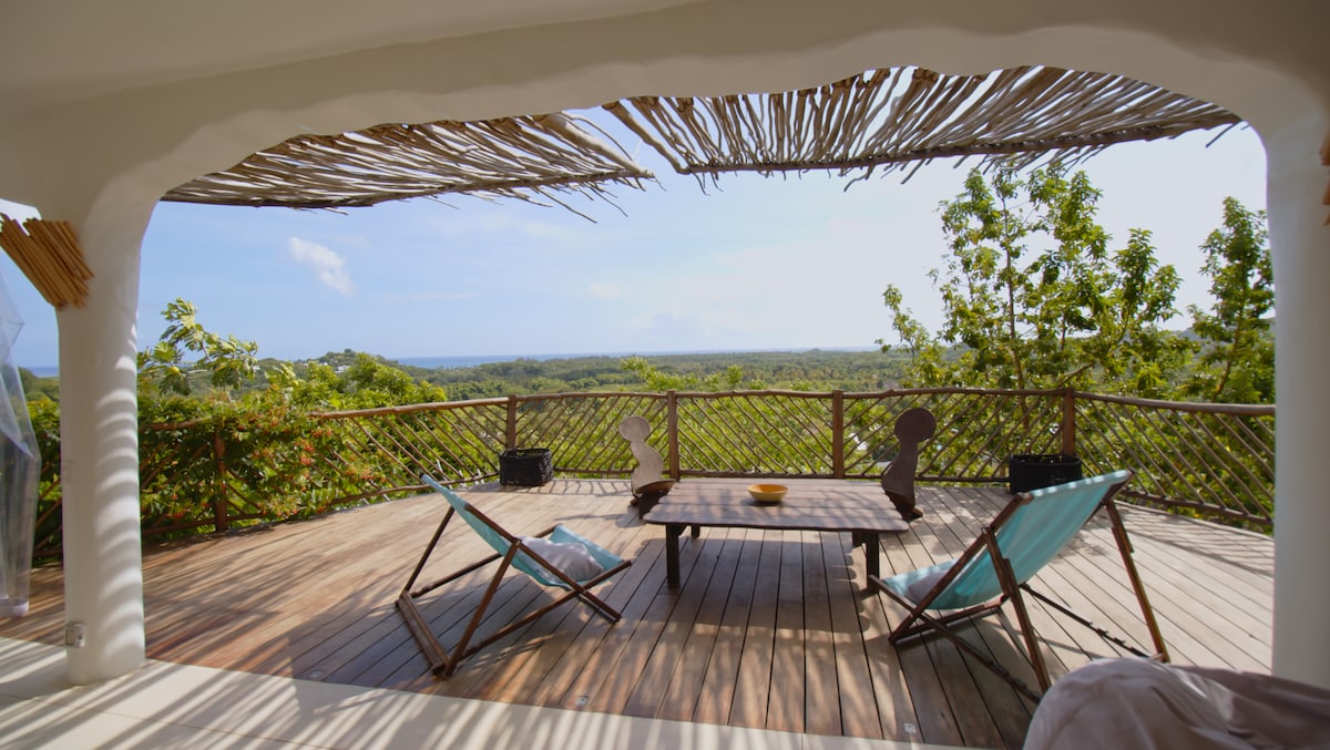 An outdoor terrace presents a serene setting with a wooden deck surrounded by lush greenery. Two turquoise lounge chairs and a wooden table are positioned to showcase the panoramic ocean view, creating a relaxing space for guests to enjoy.