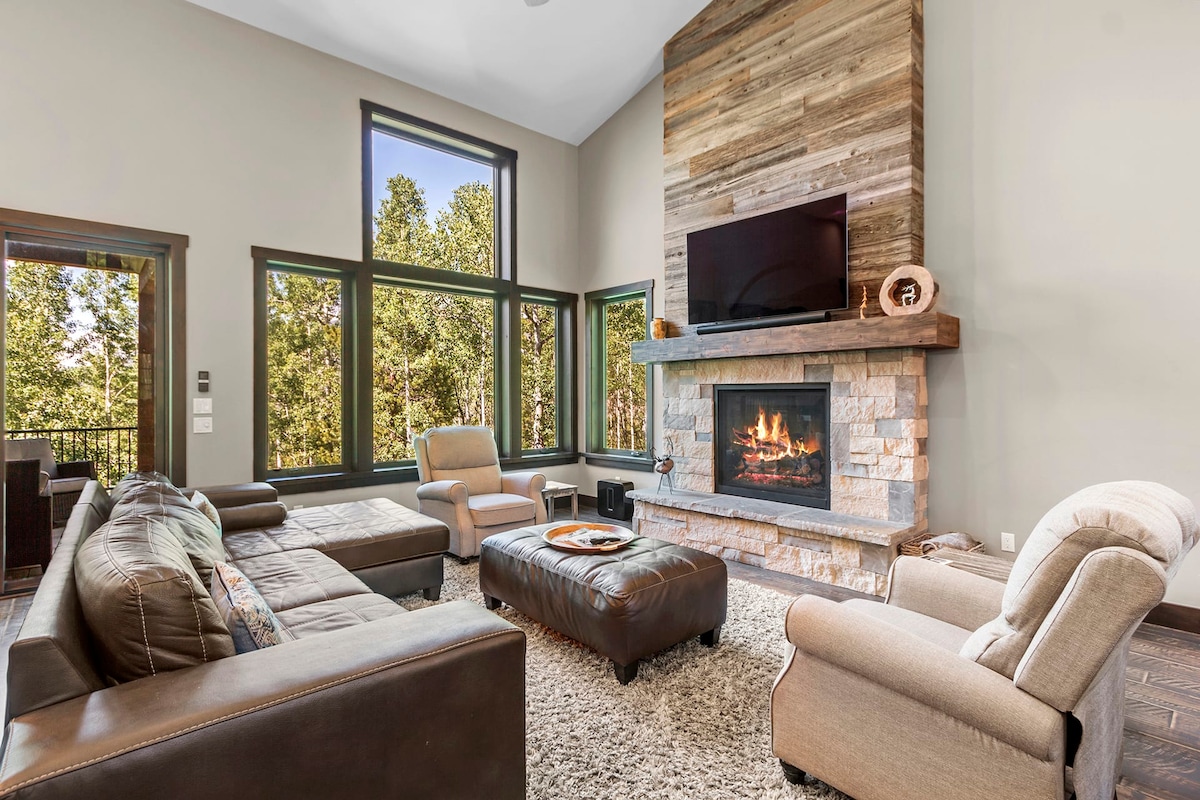 A spacious living area features large windows showcasing forested views. A modern stone fireplace is set against a wooden accent wall, while comfortable seating options surround a coffee table. Soft textures are provided by a plush area rug, enhancing the inviting atmosphere of the space.