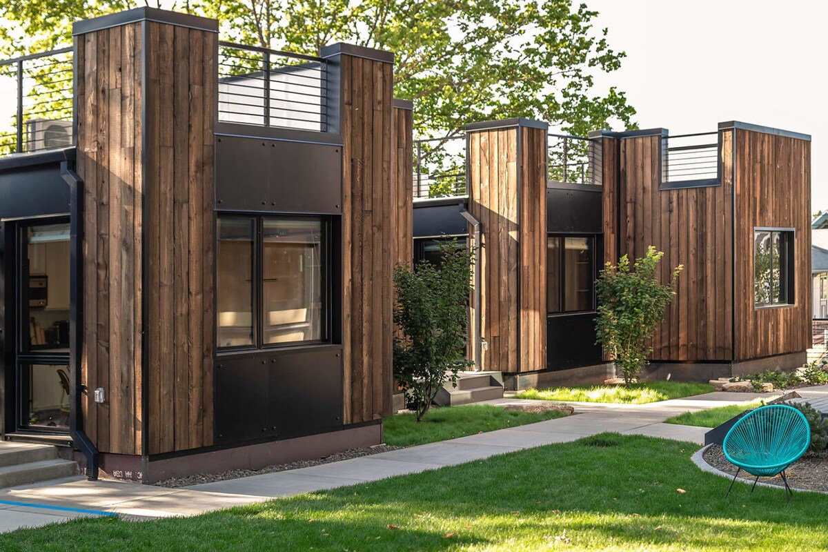 Two eco-friendly micro-studios are framed by lush green grass and a well-maintained pathway. Natural wood siding contrasts with sleek black metal accents, and large windows provide a contemporary look. A vibrant blue chair adds a pop of color to the outdoor space.