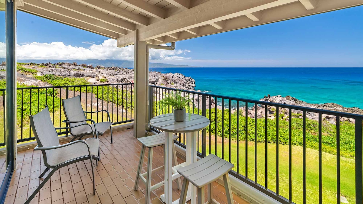 A covered lanai offers a serene spot to take in ocean views. Two lounge chairs and a small table are positioned for relaxation, with vibrant greenery and rocky shoreline visible in the foreground and the expansive blue ocean beyond.