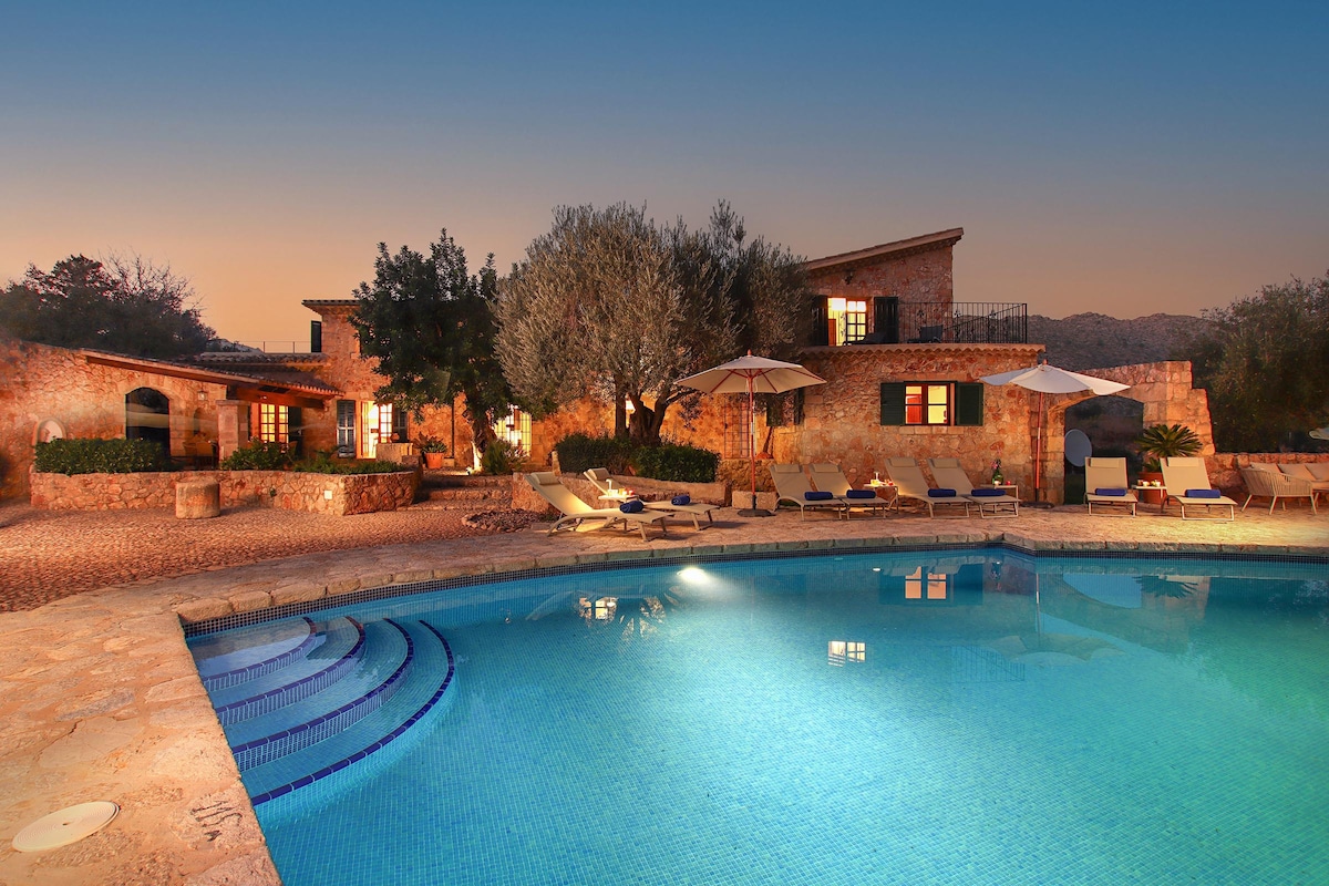 An outdoor area features a large, private swimming pool surrounded by sun loungers and shaded by umbrellas. The villa, constructed with natural stone, exhibits warm lighting that enhances its rustic charm. Lush greenery is visible in the background, contributing to a serene atmosphere.