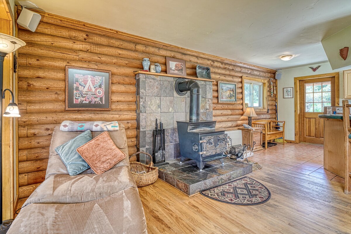 A cozy living area features a stone fireplace with a wood stove, complemented by wooden walls and warm lighting. A comfortable chaise lounge with decorative pillows is positioned nearby. A rustic rug adds texture to the wooden floor, with windows allowing natural light to enter.