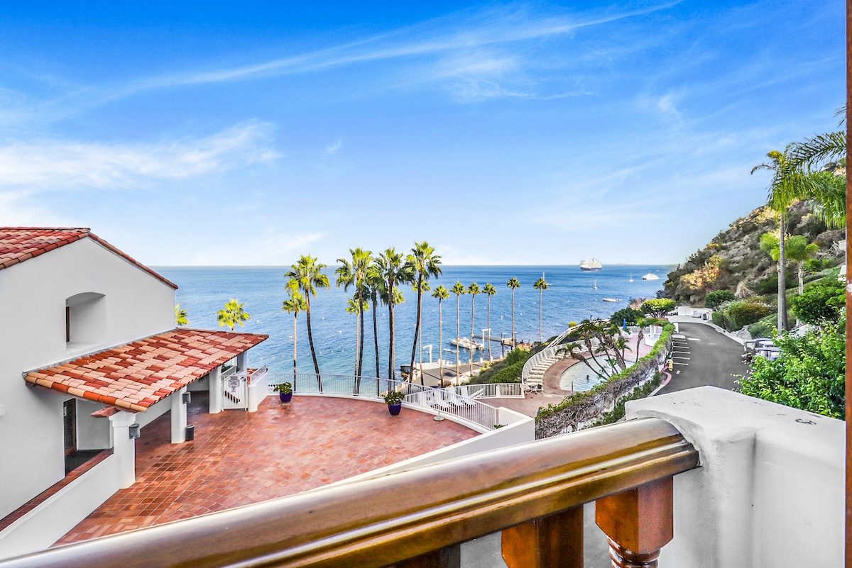 A view from the balcony showcases the stunning ocean and colorful coastline. Palm trees line the shore, with boats gently bobbing on the water. The terracotta-tiled roof complements the white villa structure, while the clear blue sky enhances the tranquil setting.