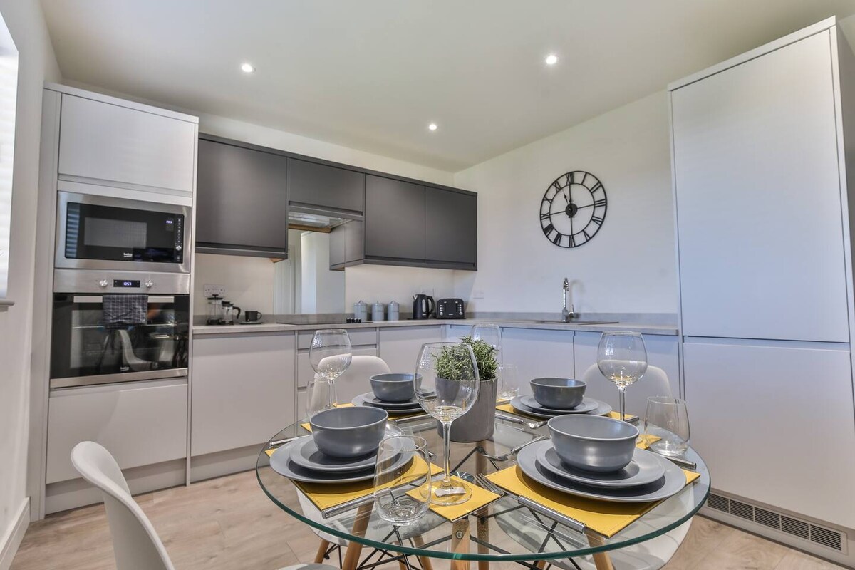 The modern kitchen features sleek cabinetry and a circular glass dining table set for six. Grey plates, wine glasses, and decorative centerpieces are arranged elegantly. Stainless steel appliances, including a microwave and kettle, are positioned strategically within the contemporary space.