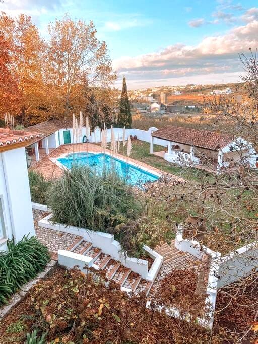 An outdoor area showcases a swimming pool surrounded by landscaped gardens. Autumn foliage in warm tones can be seen, along with a staircase leading to the pool. The scenery features a mix of greenery and earthy colors, with distant views of the landscape under a partly cloudy sky.