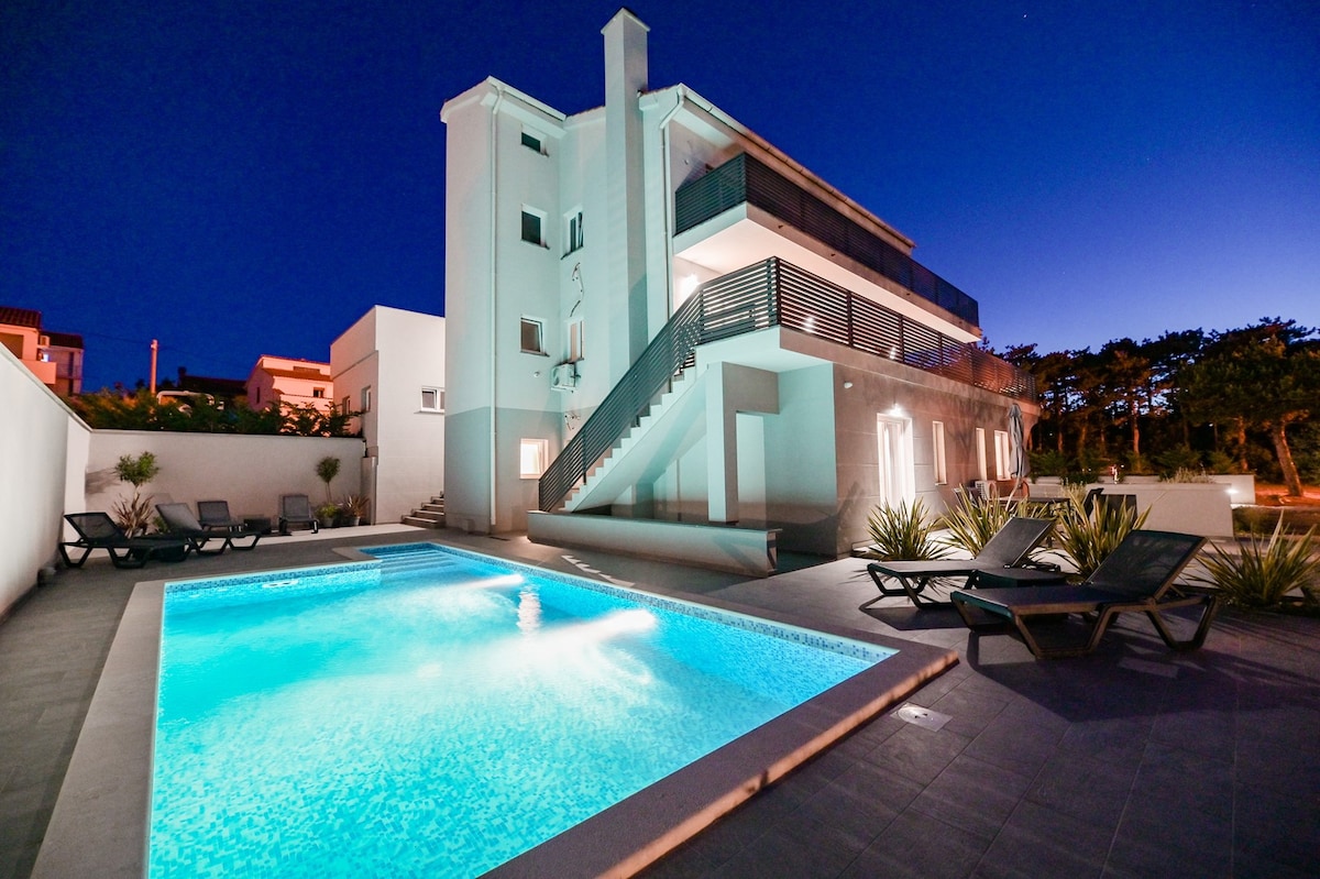 The exterior of the luxury villa is illuminated at dusk, showcasing a modern design. A sparkling pool is surrounded by lounge chairs, and the structure features multiple levels with balconies. Lush greenery is present along the property, contributing to a tranquil environment.