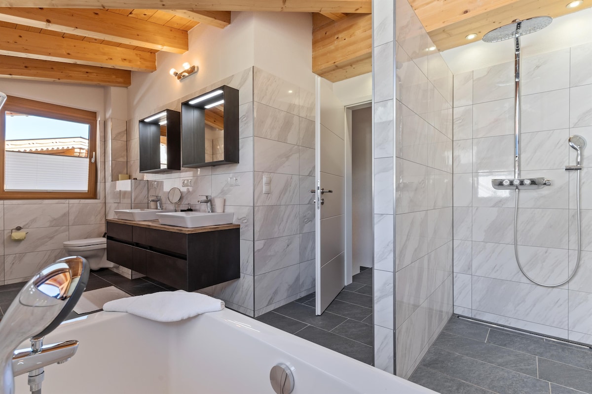 A modern bathroom is featured with a spacious shower and a luxurious bathtub. Dual sinks are positioned with stylish mirrors above. Natural light enters through a window, and sleek tiles cover the floor and walls, creating a clean and contemporary atmosphere.