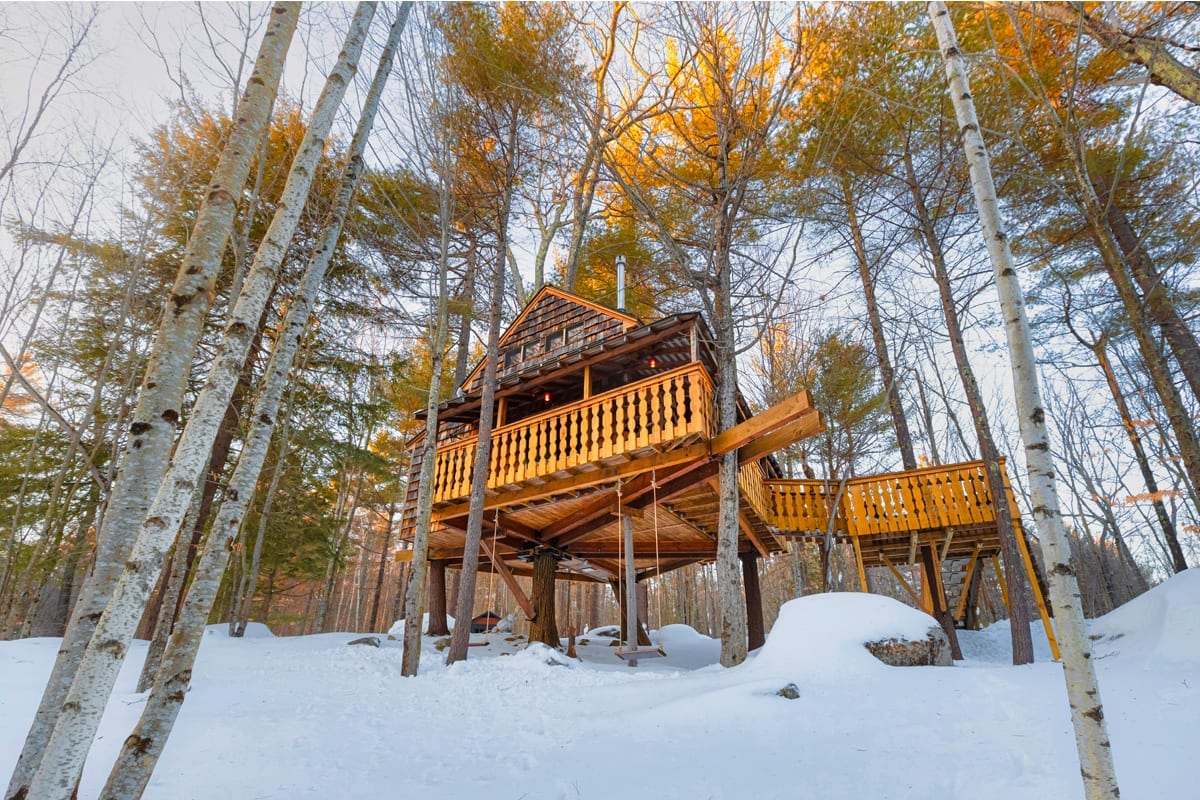 The treehouse is elevated above the snow-covered ground, supported by sturdy wooden pillars. Its rustic wooden structure features a wrap-around balcony with decorative railings, surrounded by tall trees that catch the warm glow of the setting sun.