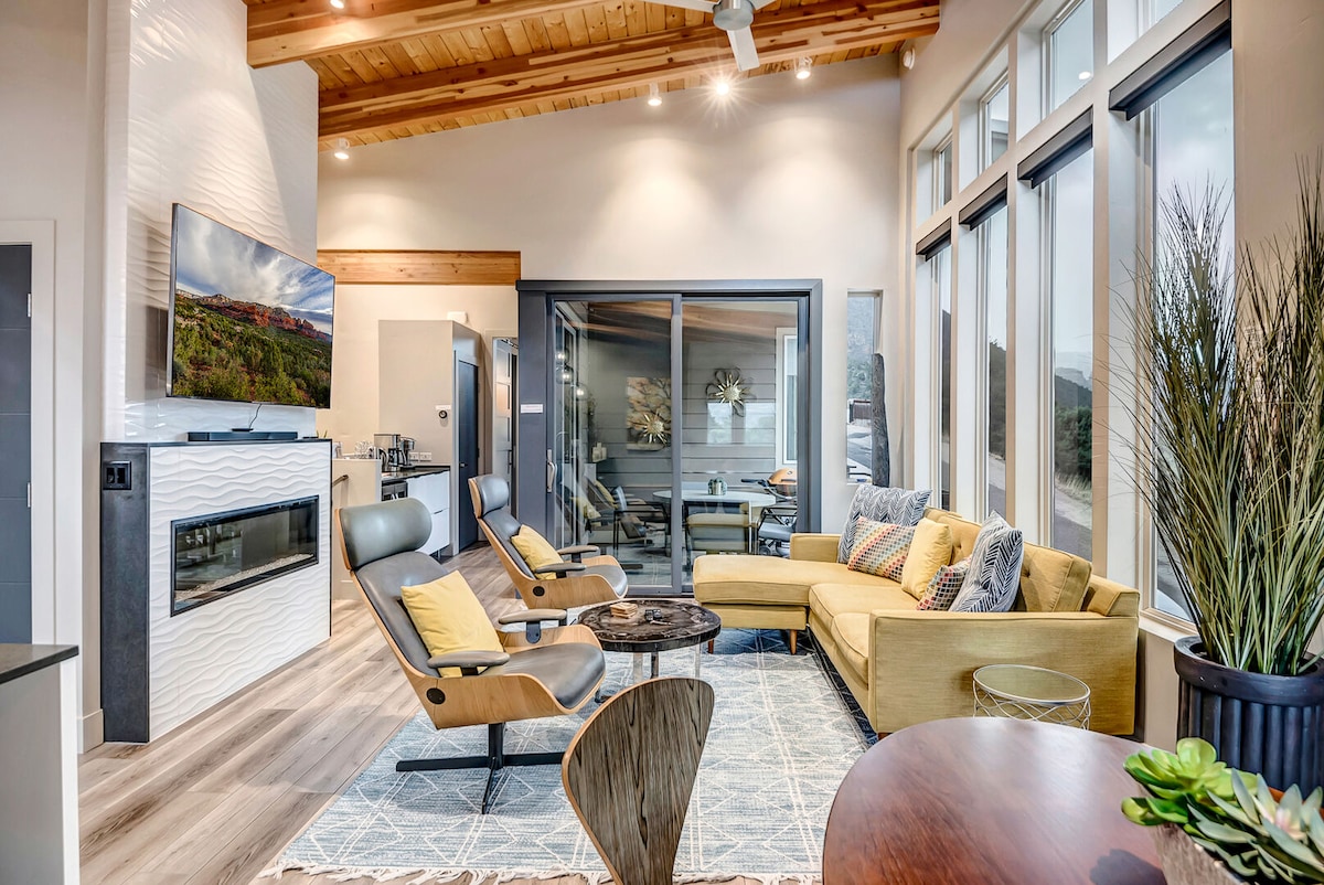 The modern living area features a yellow sectional sofa and stylish armchairs arranged around a circular coffee table. A wall-mounted Smart TV is positioned above the gas fireplace. Large windows provide ample natural light and views of the surrounding landscape, while a sliding door offers access to an outdoor space.