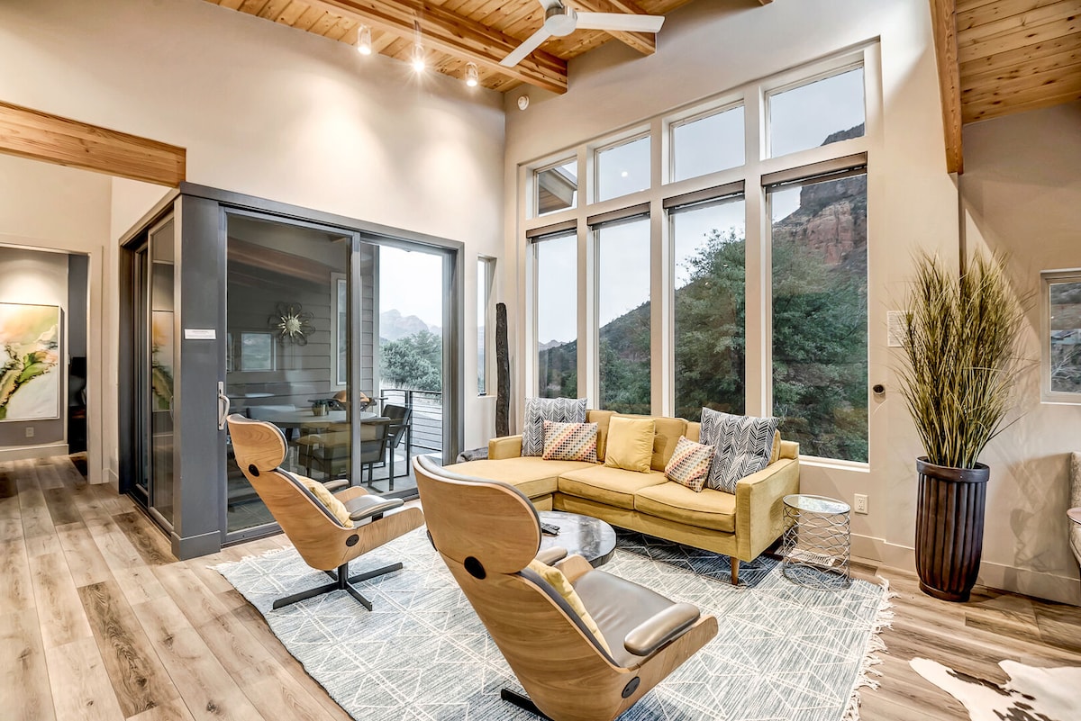 A modern living room features a yellow sectional sofa and two stylish chairs. Large windows provide views of the surrounding landscape and natural light. The space is decorated with a textured area rug and is complemented by wooden ceiling beams.