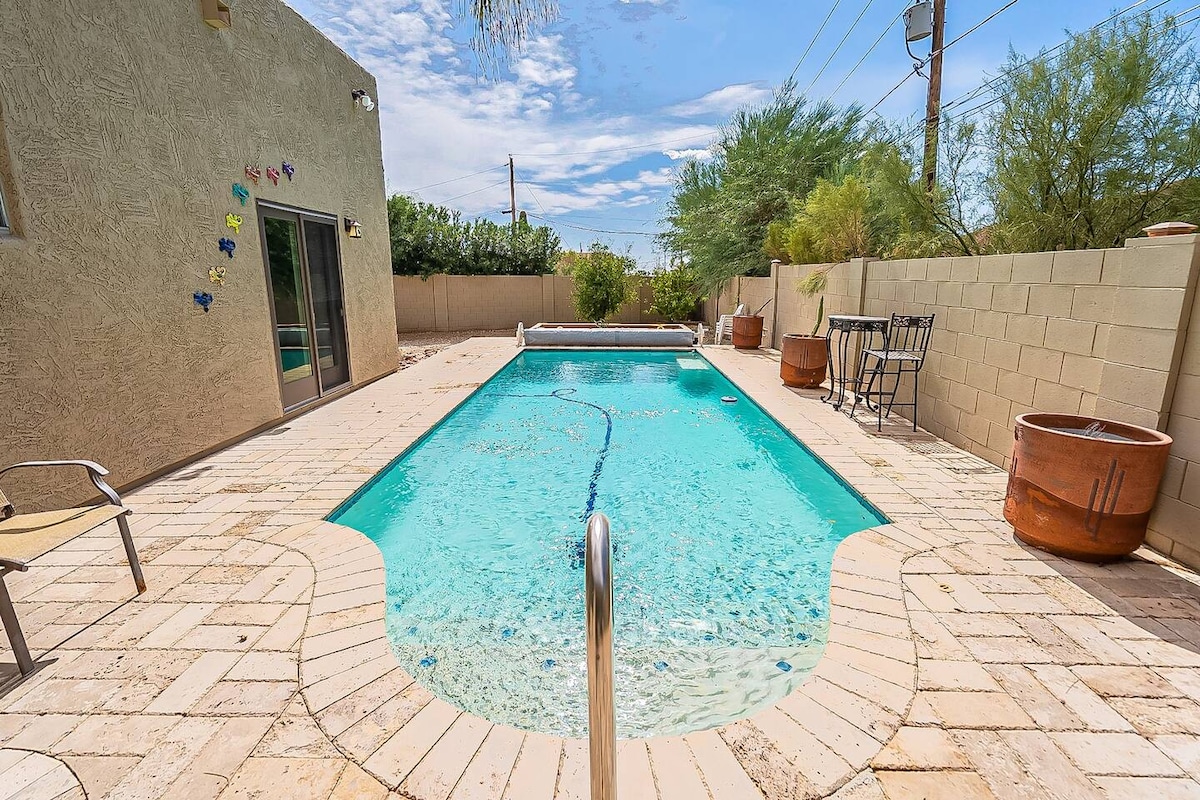 A heated pool is shown surrounded by a spacious patio, featuring a light-colored stone surface. Two wooden barrels and a seating area with a small table and chairs are positioned beside the pool. Lush greenery is visible in the background under a bright blue sky.